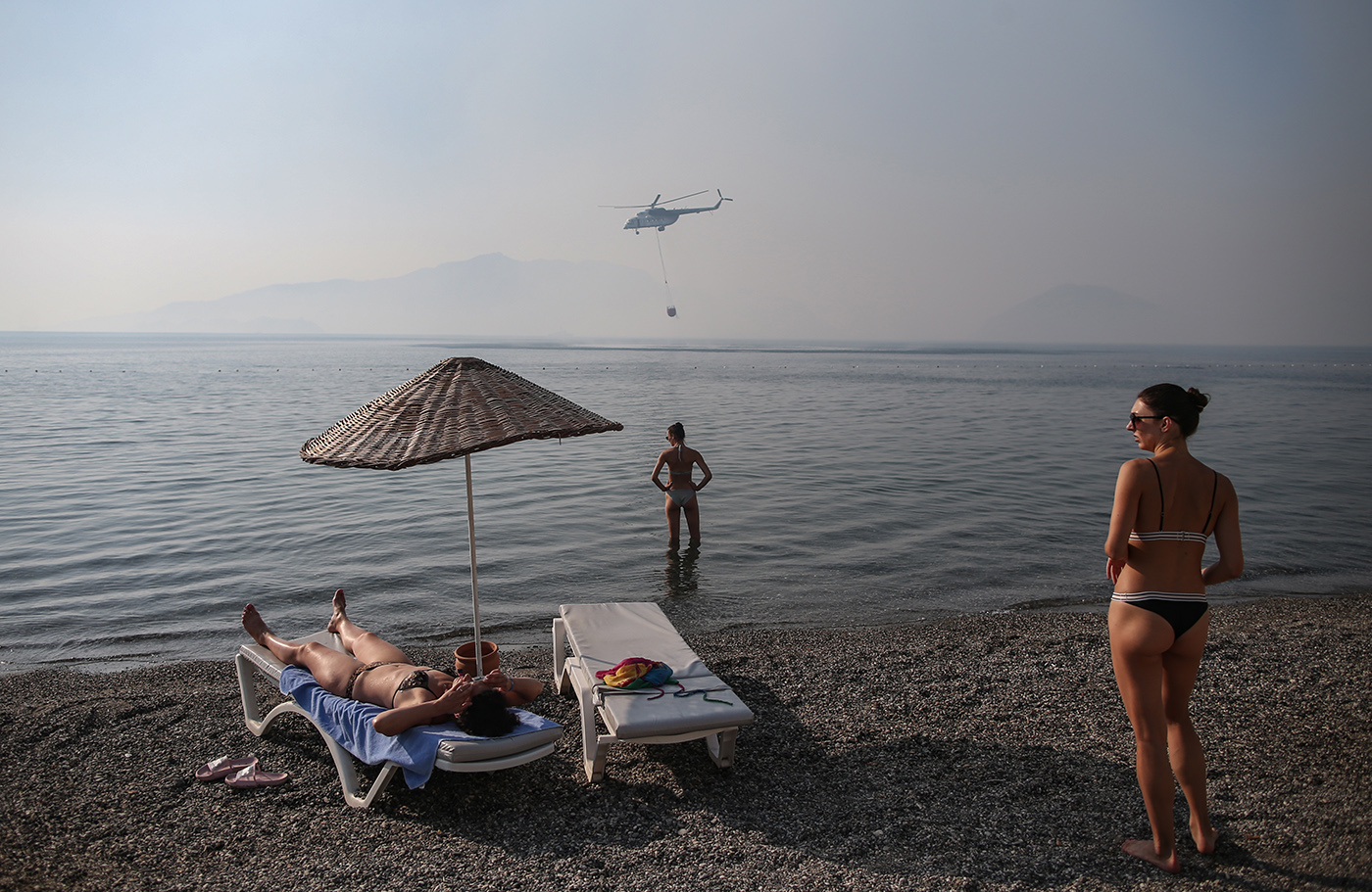 A helicopter get water from sea to put out the wildfires as tourists enjoy near the sea Marmaris district of Mugla, Turkey, 31 July 2021. According to a statement by the Turkish government